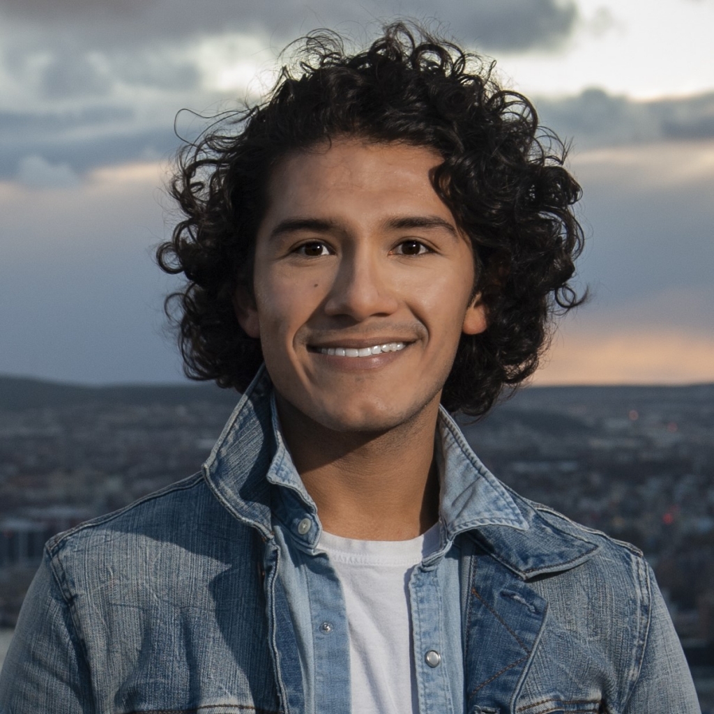 [ID: Santiago, a man with mid-neck length dark brown hair and brown eyes is smiling at the camera. He can be seen from the chest up and is smiling at the camera. He is wearing a jean jacket over a white shirt.]