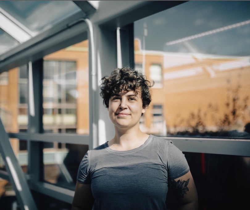 A woman with short curly brown hair and brown eyes. She is wearing a grey t-shirt and smiling at the camera. She is standing in a glass walkway with the sun shining from behind the camera.