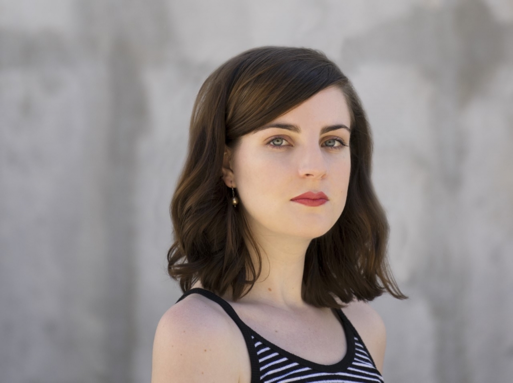 [ID: Meghan, a woman with dark brown shoulder length hair and blue eyes. She is staring at the camera and wearing a black and white striped tank top. Her background is grey.]