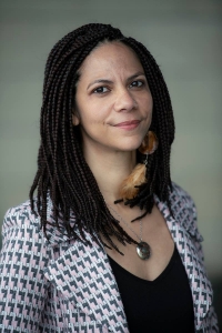 A woman with dark brown braided hair and brown eyes. She is wearing a patterned jacket over a black shirt. She has a feathered earring in her left ear and is wearing a silver necklace.