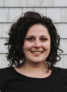 Crystal, a woman with shoulder length curly black hair. She has brown eyes and is smiling widely at the camera. She is wearing a black shirt and standing in from of a white tiled wall.