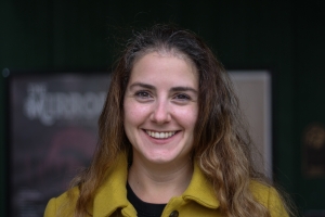 Laura, a woman with long brown hair and brown eyes is smiling with her teeth showing. She is wearing a yellow wool coat with a black shirt underneath. 