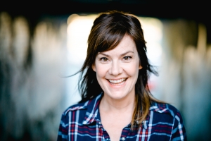 A woman with brown hair and brown eyes. She is smiling at the camera and showing her teeth. She is wearing a blue and red plaid shirt.