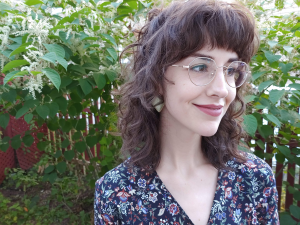 Sara, a woman with shoulder length brown curly hair is looking to the side of the camera. She has brown eyes and round wire rim glasses. She is wearing a patterned V-neck shirt and standing in front of greenery..