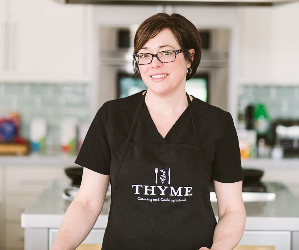 Jennifer, a owman with short straight brown hair and brown eyes is smiling at the camera. She is standing in a kitchen and wearing a black shirt with a black apron over top that says Thyme Catering and Cooking School.