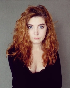 Alison, a woman with long wavy red hair and blue eyes is facing the camera. She has a nose ring and is wearing a black long sleeve shirt.