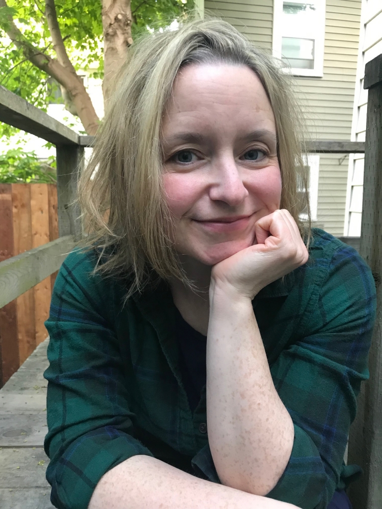 Willow, a woman with shoulder length blonde hair and blue eyes is smiling at the camera, resting her chin on her left hand. She is wearing a dark green and blue plaid shirt. She is sitting on a deck with a tree behind her.