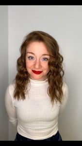 Allison, a person with long wavy brown hair and blue eyes is wearing bright red lipstick and a white turtleneck. They stand in front of a white wall. 