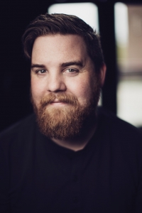 Brian Kenny, a man with short brown hair and a brown/blonde beard is wearing a black shirt. A blurry window is in the background.