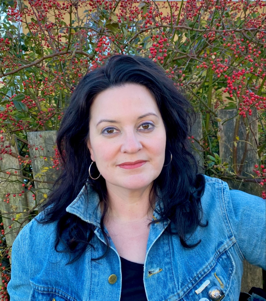 Nicole, a woman with wavy black hair past her shoulders is wearing a blue jean jacket over a black shirt. She has silvery hoop earrings and pink lipstick. She stands in front of a dogberry tree.