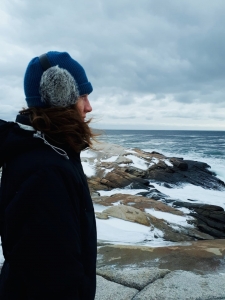 Kaitlin, a woman with long brown hair, stares out towards the ocean. She is wearing a blue hat with grey furry ear muffs over it and a dark blue coat. Rocks and the ocean are in her background.