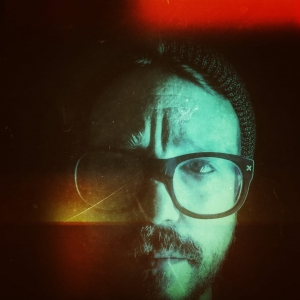 tyler, a man with glasses and a beard is shown from neck up. He is wearing a hat. The filter on the picture makes the color pale blue. Red can be seen in the top right corner and yellow in the bottom left.