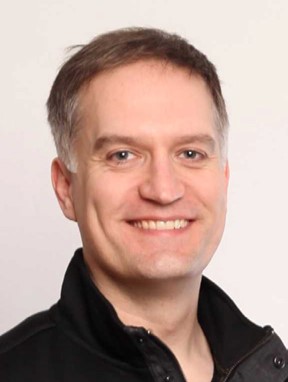 Aiden, a man with short brown and grey hair is smiling at the camera. He is wearing a black collared shirt.