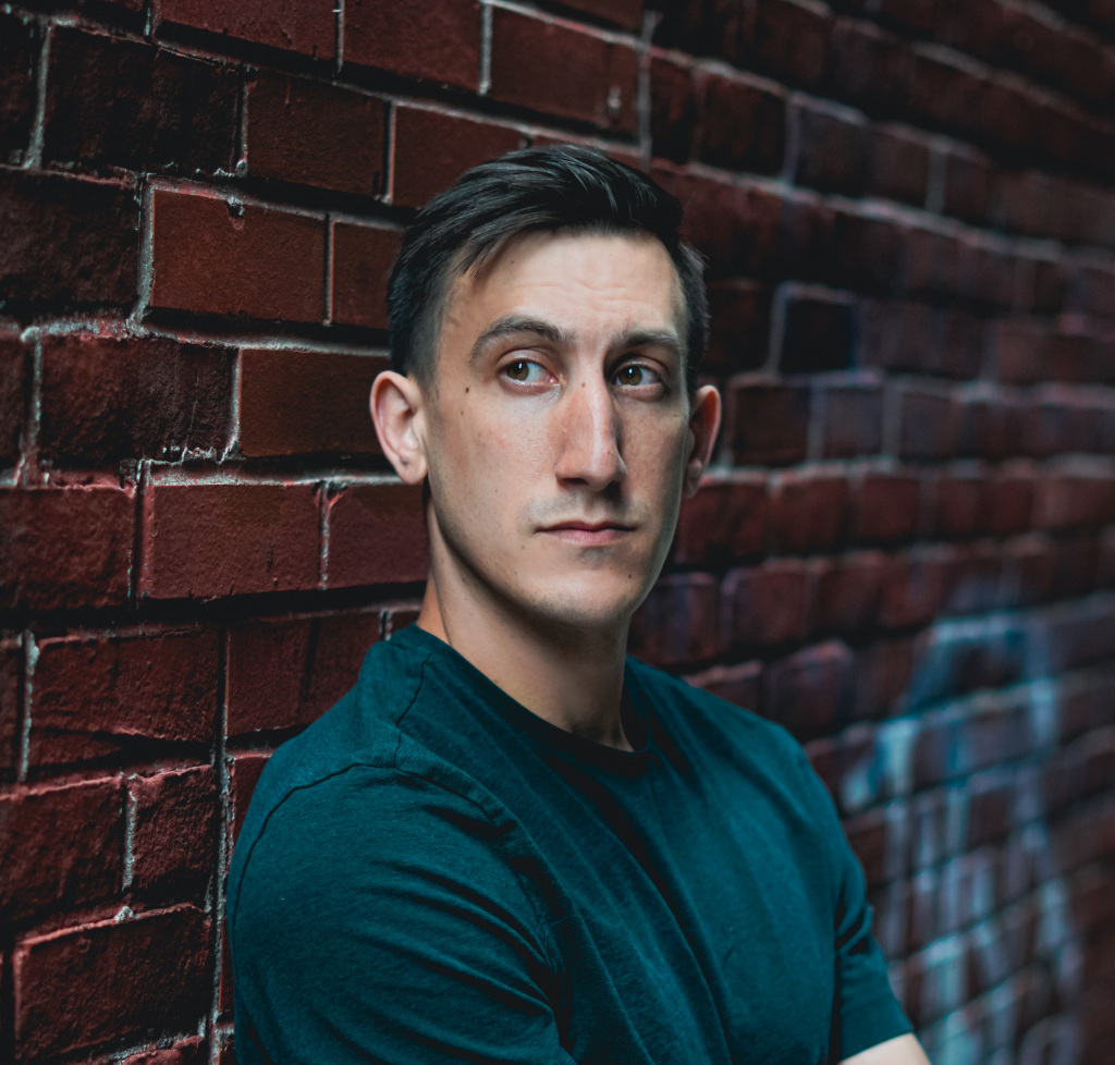 A photo of Darren, a white man with short black hair and brown eyes. He leans against a brick wall and is wearing a blue t-shirt.