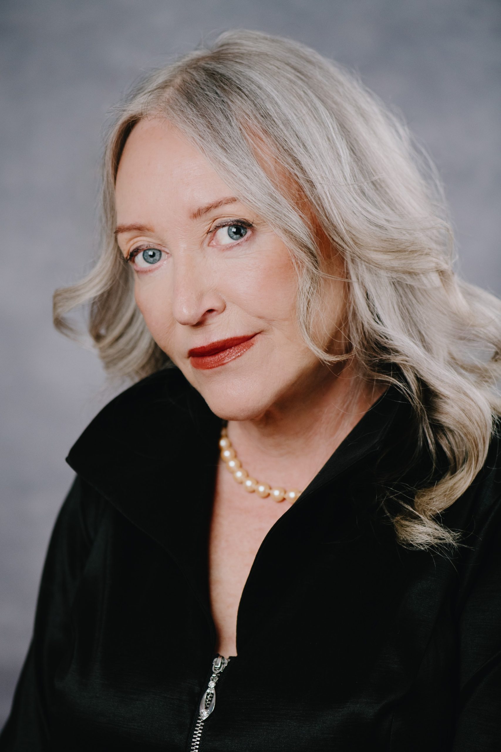 [ID: A colour photo of a white women with long grey hair. She is wearing a black blouse and a string of pearls around her neck. She is smiling and looking at the camera.]