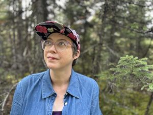 [ID: A woman wearing glasses, a blue collared shirt, and a baseball hat with flower pattern. She has short brown hair and is looking off to the side, smiling. There are pine trees in the background.]
